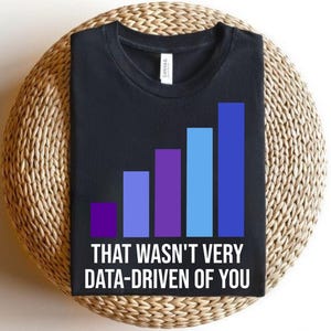 以下が含まれることがあります： 紫と青の色合いのデータ視覚化グラフィックが特徴の黒いTシャツ。「THAT WASN'T VERY DATA-DRIVEN OF YOU」というテキストが下に印刷されています。Tシャツは、丸い織りの表面に表示されています。