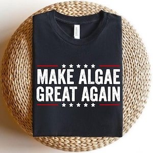 May include: A black short-sleeved t-shirt featuring the white distressed text "MAKE ALGAE GREAT AGAIN" centered on the chest. The design includes two horizontal red lines above and below the text, with five white stars positioned between the lines.