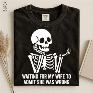 May include: A black crew neck t-shirt featuring a white graphic of a skeleton resting its chin on one hand with the text "WAITING FOR MY WIFE TO ADMIT SHE WAS WRONG" printed in white capital letters below the skeleton illustration.