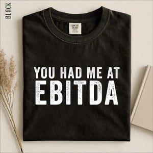 May include: A black short-sleeved t-shirt with a crew neckline. The front of the shirt features the white distressed-style text "YOU HAD ME AT EBITDA" printed in a bold, sans-serif font. The garment includes a Comfort Colors brand neck label.