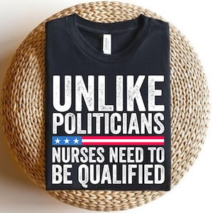 Puede incluir: Camiseta negra con texto blanco: "UNLIKE POLITICIANS NURSES NEED TO BE QUALIFIED". El diseño incluye un gráfico rojo, blanco y azul. La camiseta se muestra sobre una superficie circular tejida.