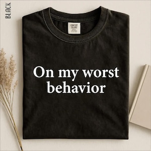 May include: A black crew neck t-shirt featuring the white text On my worst behavior printed across the chest. The shirt has a Comfort Colors brand label on the inside of the collar and is displayed flat with a neutral background.