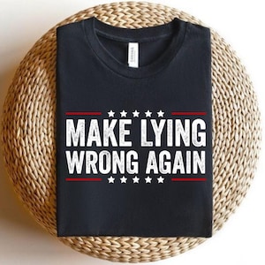 May include: Black t-shirt with the text "MAKE LYING WRONG AGAIN" in white, with red accents and star details. The shirt is displayed on a woven, circular surface.