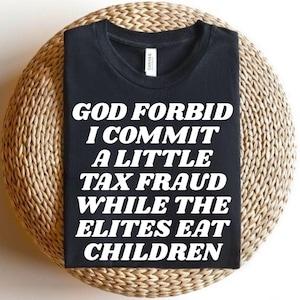 Op de afbeelding: Een zwart t-shirt met korte mouwen en de witte tekst "GOD FORBID I COMMIT A LITTLE TAX FRAUD WHILE THE ELITES EAT CHILDREN" gedrukt in een vet, schreefloos lettertype in het midden van de borst van het kledingstuk.