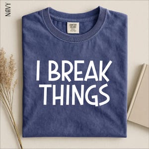 May include: A navy blue short-sleeved t-shirt with a crew neckline. The shirt features the white text "I BREAK THINGS" printed in a bold, sans-serif font across the center. A Comfort Colors brand label is visible on the inside of the collar.