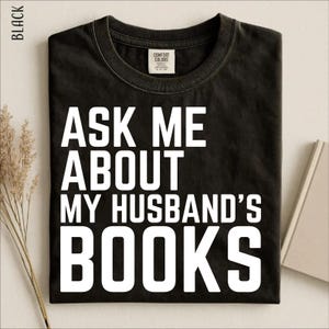 Könnte beinhalten: Ein schwarzes kurzärmeliges T-Shirt mit weißem Text auf der Vorderseite, der "ASK ME ABOUT MY HUSBAND'S BOOKS" lautet. Das T-Shirt hat einen Rundhalsausschnitt und ein Markenetikett von Comfort Colors an der Innenseite des Kragens.