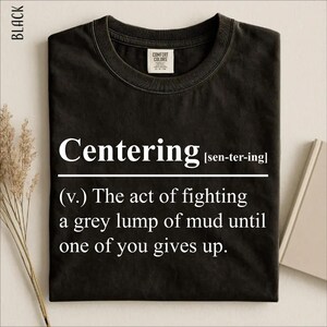 Könnte beinhalten: Ein schwarzes kurzärmeliges T-Shirt mit weißem Text, der lautet Centering [sen-ter-ing] (v.) The act of fighting a grey lump of mud until one of you gives up. Das T-Shirt hat einen Rundhalsausschnitt und ein Comfort Colors Markenetikett am Innenkragen.