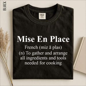Puede incluir: Una camiseta negra de cuello redondo con texto en blanco que dice: Mise En Place, French (miz ã plas), (n) To gather and arrange all ingredients and tools needed for cooking. La camiseta tiene una etiqueta de la marca Comfort Colors.