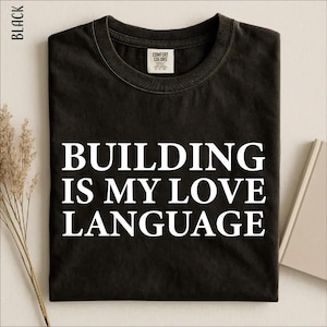 May include: A black short-sleeved t-shirt featuring the white text "BUILDING IS MY LOVE LANGUAGE" printed in a bold, serif font across the chest. The garment has a crew neckline and a Comfort Colors brand label visible inside the collar.