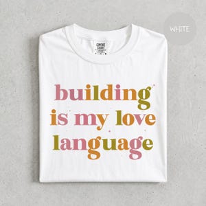 Pode incluir: Uma t-shirt branca de manga curta com o texto "building is my love language" impresso numa fonte serifada multicolorida com letras cor-de-rosa, laranja e verde-azeitona. Pequenos ícones de estrelas estão espalhados pelo texto na frente.