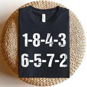 Könnte beinhalten: Schwarzes T-Shirt mit weißen, verwaschenen Zahlen in zwei Reihen: 1-8-4-3 und 6-5-7-2. Das T-Shirt wird auf einer runden, gewebten, hellbraunen Oberfläche präsentiert.