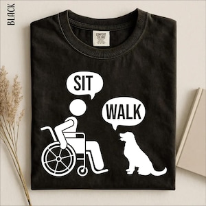 May include: A black t-shirt featuring a white graphic of a person in a wheelchair with a speech bubble that says SIT and a dog with a speech bubble that says WALK. The shirt has a crew neckline and a Comfort Colors brand label at the inner collar.