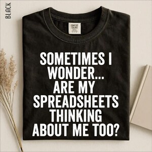 Può includere: Una t-shirt nera a girocollo con il testo bianco dall'aspetto usurato: SOMETIMES I WONDER... ARE MY SPREADSHEETS THINKING ABOUT ME TOO? La maglietta presenta un'etichetta del marchio Comfort Colors all'interno del colletto ed è disposta in piano.