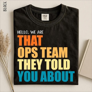 May include: A black short-sleeve t-shirt with a crew neckline. The graphic print features the text HELLO, WE ARE THAT OPS TEAM THEY TOLD YOU ABOUT in stacked lines of orange, yellow, and light blue, with a Comfort Colors brand label at the neck.