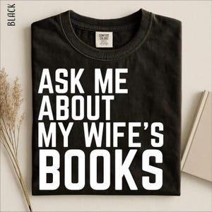 Könnte beinhalten: Ein schwarzes Kurzarm-T-Shirt mit Rundhalsausschnitt und dem weißen Schriftzug "ASK ME ABOUT MY WIFE'S BOOKS" in fetter, serifenloser Schrift auf der Brust. Das T-Shirt ist mit einem Comfort Colors Markenetikett am Kragen versehen.