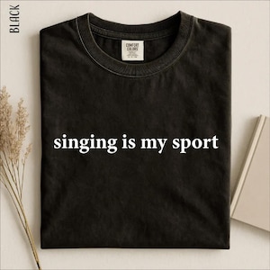 Puede incluir: Una camiseta negra de cuello redondo con el texto blanco "singing is my sport" impreso en el centro. La prenda tiene un cuello acanalado y una etiqueta de la marca Comfort Colors visible en el interior del escote, con un diseño sencillo.