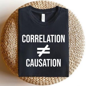 May include: Black t-shirt with the white text "CORRELATION ≠ CAUSATION". The shirt is displayed on a woven, circular surface. The text is in a bold, sans-serif font.