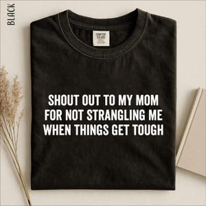 May include: A black crew neck t-shirt featuring white text that reads, "SHOUT OUT TO MY MOM FOR NOT STRANGLING ME WHEN THINGS GET TOUGH." The shirt has a Comfort Colors brand label on the inside of the collar and is displayed flat.