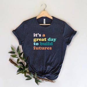 Peut inclure: T-shirt bleu marine avec l'inscription "it's a great day to build futures" en lettres color&eacute;es. Le t-shirt est suspendu &agrave; un cintre en bois sur fond blanc. Une branche avec des feuilles vertes est plac&eacute;e en bas &agrave; gauche.