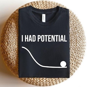 Peut inclure: T-shirt noir avec le texte blanc "I HAD POTENTIAL" au-dessus d'un graphique blanc représentant une rampe incurvée et une boule. Le t-shirt est présenté sur une surface circulaire tissée.
