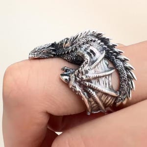 Peut inclure: Une bague argentée en forme de dragon. La bague présente un design de dragon détaillé, avec la tête et la queue du dragon enroulées autour du doigt. Les ailes du dragon sont visibles sur le côté de la bague.