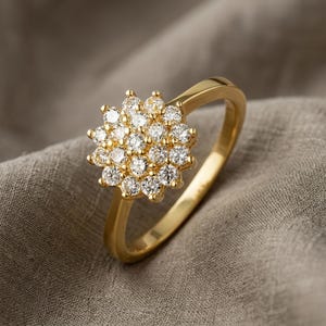May include: A gold ring featuring a cluster of small, round, clear gemstones arranged in a floral pattern. The ring's band is a polished gold color, and the gemstones sparkle against the neutral background.
