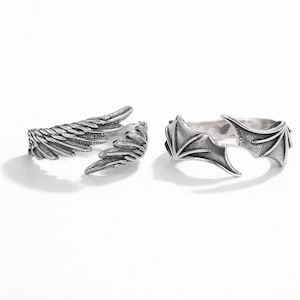 May include: Two silver rings. One ring features a detailed wing design, while the other showcases a bat wing design. Both rings are open-ended, allowing for adjustable sizing. The rings are set against a white background.