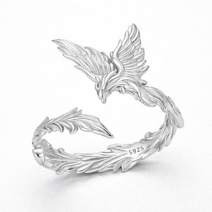 Könnte beinhalten: Ein silberner Ring in Form eines Phönix. Der Ring weist detaillierte Federdesigns auf, und der Phönix wird mit ausgebreiteten Flügeln dargestellt. Der Ring ist mit "S925" markiert.