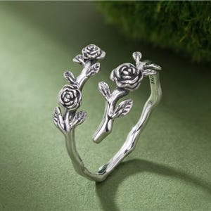 Peut inclure: Bague argentée avec un motif floral. La bague présente trois fleurs de roses et des feuilles sur des branches qui entourent le doigt. La bague est posée sur un fond vert.