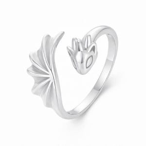 Puede incluir: Un anillo plateado con un diseño de dragón. El anillo presenta una cabeza y un ala de dragón. La cabeza del dragón está en un extremo del anillo y el ala en el otro. El anillo es de metal brillante.