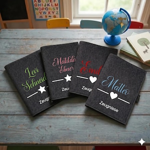 Könnte beinhalten: Vier dunkelgraue Filznotizbücher mit personalisierten Namen in verschiedenen Farben. Jedes Notizbuch hat einen Namen, eine Stern- oder Herzgrafik und das Wort "Zeugnisse" oder "Zeugr" in Weiß. Ein Globus und Bücher sind im Hintergrund.