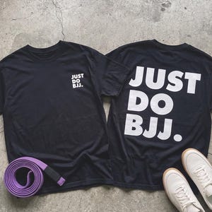 May include: Two black short-sleeve t-shirts featuring the white text "JUST DO BJJ." on the front and a larger version on the back. A coiled purple martial arts belt with a black tip and a pair of white trainers are placed near the shirts.