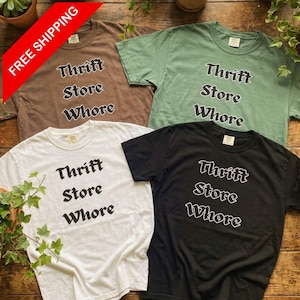 May include: Four t-shirts in brown, green, white, and black, each with the text "Thrift Store Whore" in a bold, stylized font. The shirts are displayed on a wooden surface, with a "FREE SHIPPING" banner in the upper left corner.