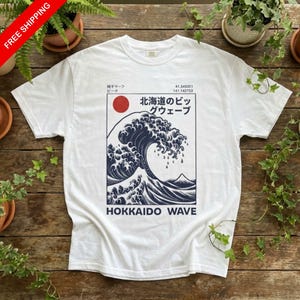 May include: White t-shirt with a navy blue and white graphic of a large wave, a red sun, and Japanese text. The text "HOKKAIDO WAVE" is printed below the wave design. The shirt is displayed on a wooden surface.