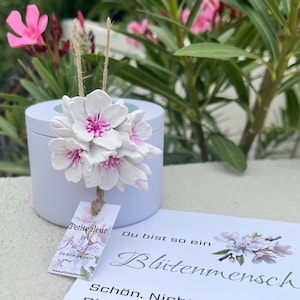 Puede incluir: Un racimo de flores de arcilla blancas y rosas unidas a un cordel de yute, apoyado contra un recipiente cilíndrico blanco. Una etiqueta de papel rectangular cuelga de las flores con el texto Petite fleur by Pfalzblüte. Una tarjeta muestra el texto Du bist so ein Blütemensch. Schön. Nicht.
