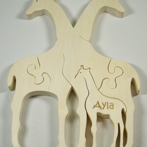 Op de afbeelding: Een houten puzzel met twee giraffen, een groter dan de andere. De kleinere giraffe heeft de naam "Ayla" gegraveerd op zijn zijkant. De puzzelstukken passen in elkaar om een ​​volledig beeld van de giraffen te vormen.