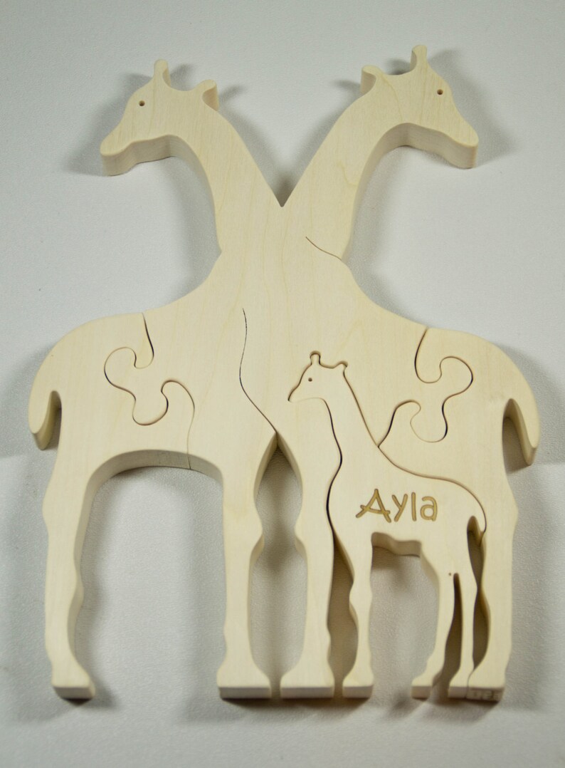 Op de afbeelding: Een houten puzzel met twee giraffen, een groter dan de andere. De kleinere giraffe heeft de naam "Ayla" gegraveerd op zijn zijkant. De puzzelstukken passen in elkaar om een ​​volledig beeld van de giraffen te vormen.