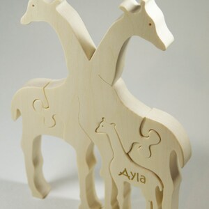 Op de afbeelding: Een houten puzzel van drie giraffen, twee volwassenen en een baby. De babygiraf heeft de naam "Ayla" gegraveerd op zijn zijkant.