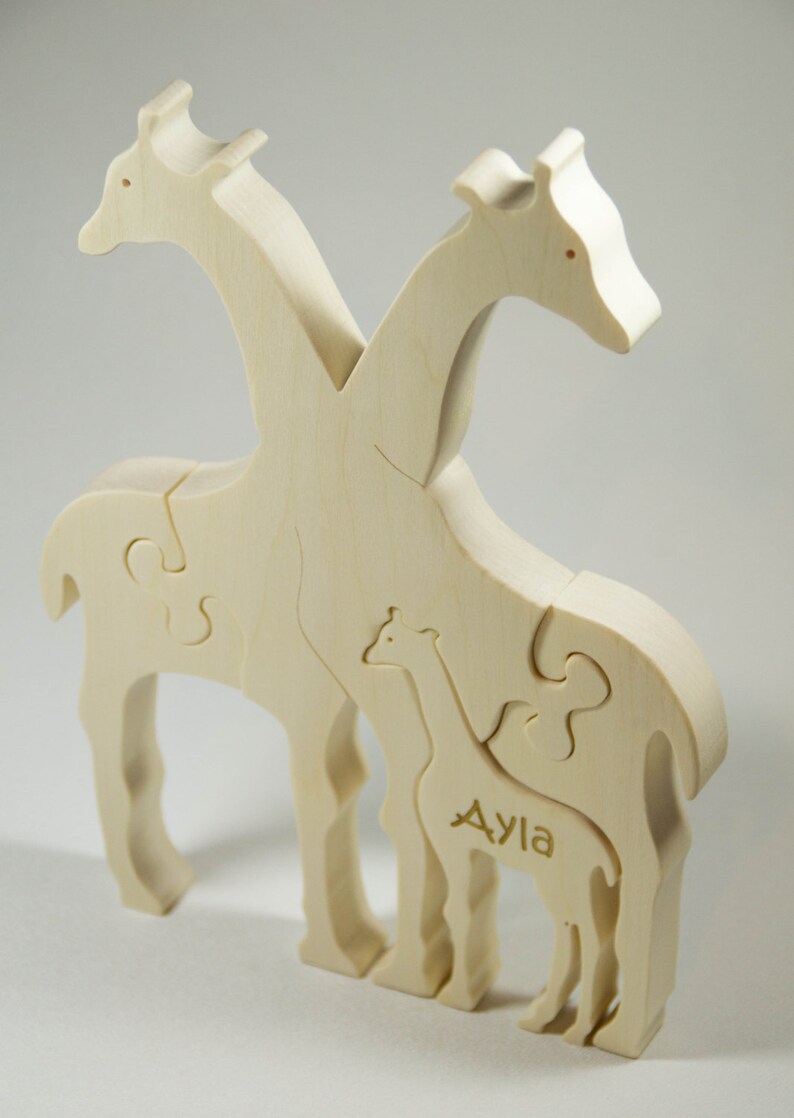Op de afbeelding: Een houten puzzel van drie giraffen, twee volwassenen en een baby. De babygiraf heeft de naam "Ayla" gegraveerd op zijn zijkant.