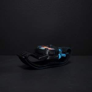 May include: A black wristwatch with a round face and a black strap. The watch face has orange and blue accents. The watch is resting on a dark surface, with a dark background.