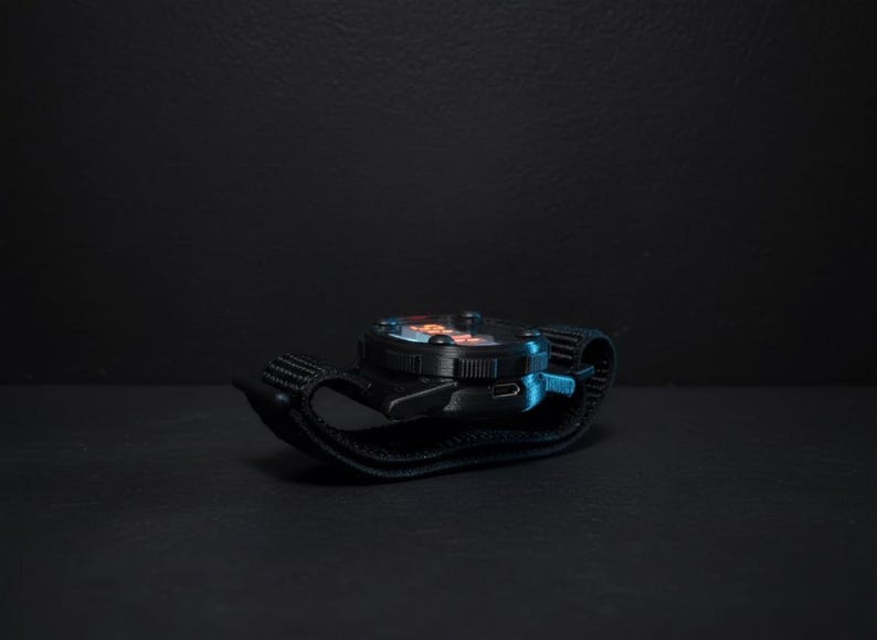 May include: A black wristwatch with a round face and a black strap. The watch face has orange and blue accents. The watch is resting on a dark surface, with a dark background.