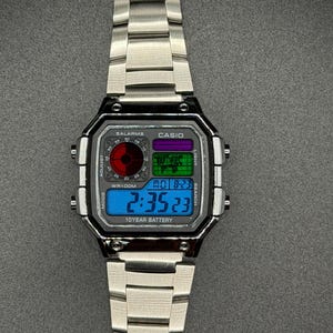 May include: A silver-toned digital wristwatch with a rectangular face and matching band. The watch face displays the time, date, and other functions in blue, green, and purple. The watch has the text "CASIO" and "SALARMS".