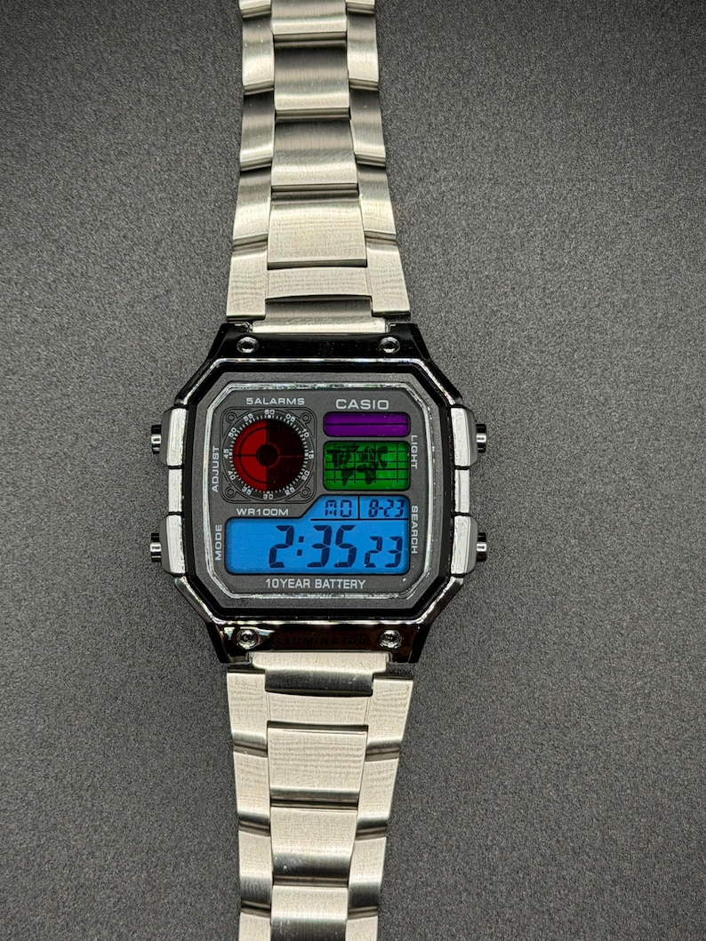 May include: A silver-toned digital wristwatch with a rectangular face and matching band. The watch face displays the time, date, and other functions in blue, green, and purple. The watch has the text "CASIO" and "SALARMS".