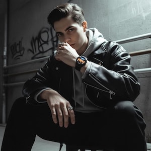 May include: A black smartwatch with a digital display is worn on a wrist. The watch has a black band and a dark face with orange and blue accents. The person is wearing a black leather jacket, a gray hoodie, and black pants.