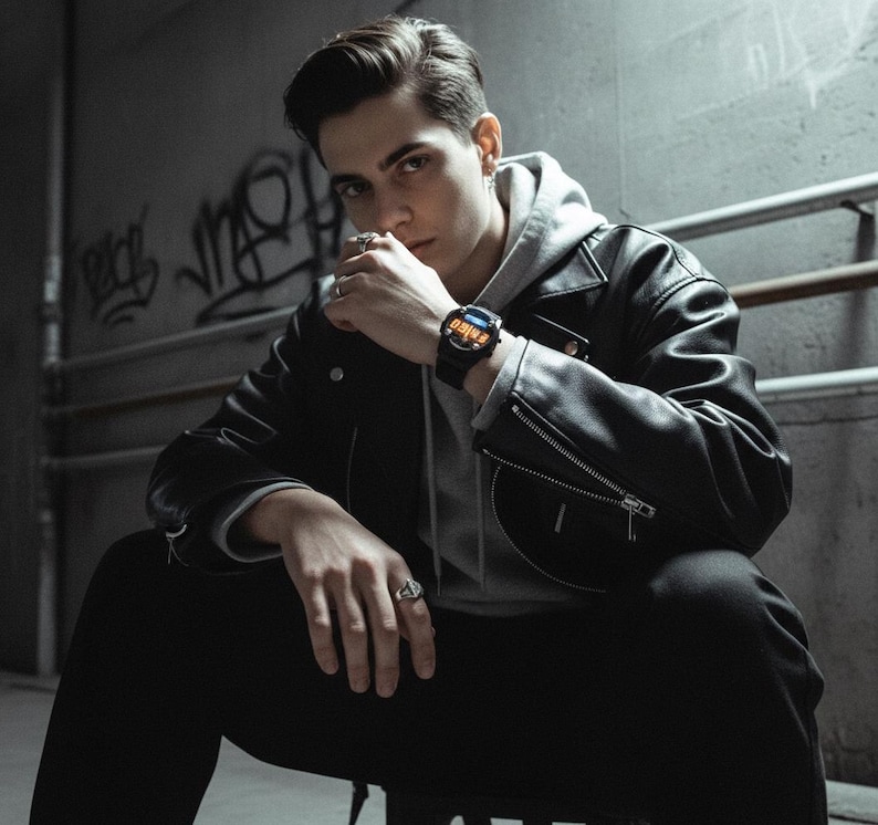 May include: A black smartwatch with a digital display is worn on a wrist. The watch has a black band and a dark face with orange and blue accents. The person is wearing a black leather jacket, a gray hoodie, and black pants.