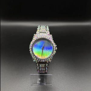 May include: A silver wristwatch with a round face and a band adorned with iridescent crystals. The watch face displays a rainbow-colored, oil-slick effect. The watch is displayed on a clear stand against a dark background.