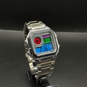 May include: A silver Casio digital watch with a stainless steel band. The watch face displays the time, date, and other functions on a blue and green digital display. The watch has the text "WORLD TIME", "CASIO", "10 YEAR BATTERY", and "ILLUMINATOR".