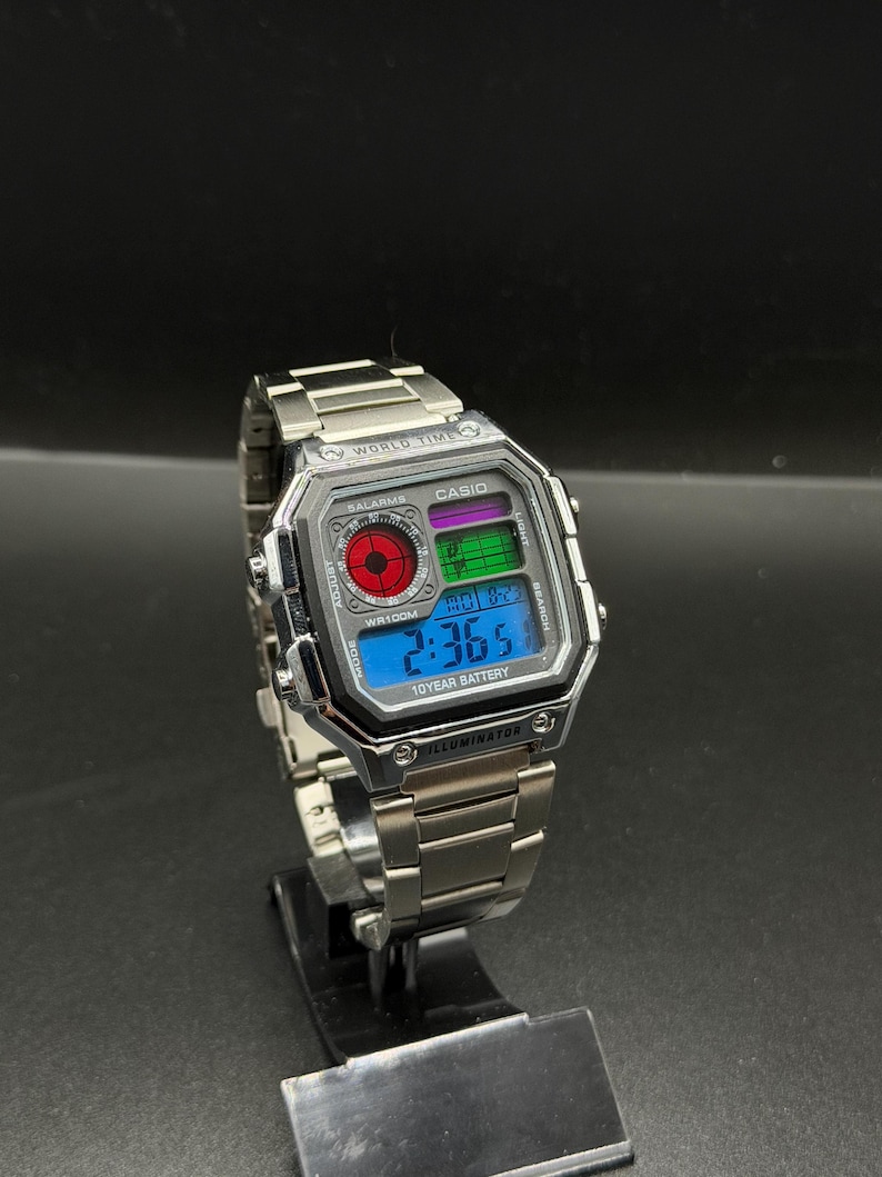 May include: A silver Casio digital watch with a stainless steel band. The watch face displays the time, date, and other functions on a blue and green digital display. The watch has the text "WORLD TIME", "CASIO", "10 YEAR BATTERY", and "ILLUMINATOR".