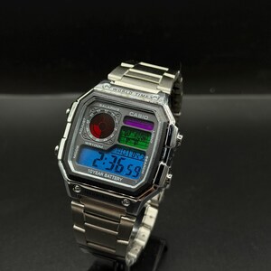 May include: A silver Casio digital watch with a rectangular face and a stainless steel band. The watch face displays the time, date, and other functions in a blue, green, and purple color scheme. The watch has the text "CASIO" and "10 YEAR BATTERY".