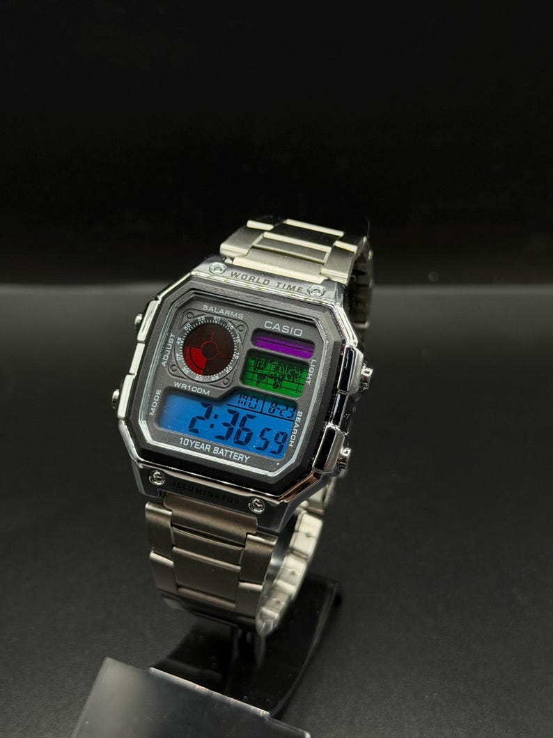 May include: A silver Casio digital watch with a rectangular face and a stainless steel band. The watch face displays the time, date, and other functions in a blue, green, and purple color scheme. The watch has the text "CASIO" and "10 YEAR BATTERY".